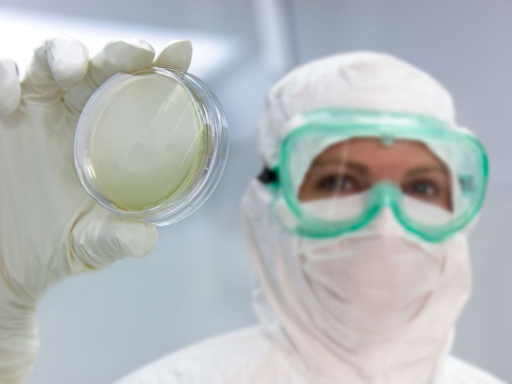 Woman wearing protective gear and googles, slightly out of focus, holding up a Petri dish with a gloved hand that is in focus.