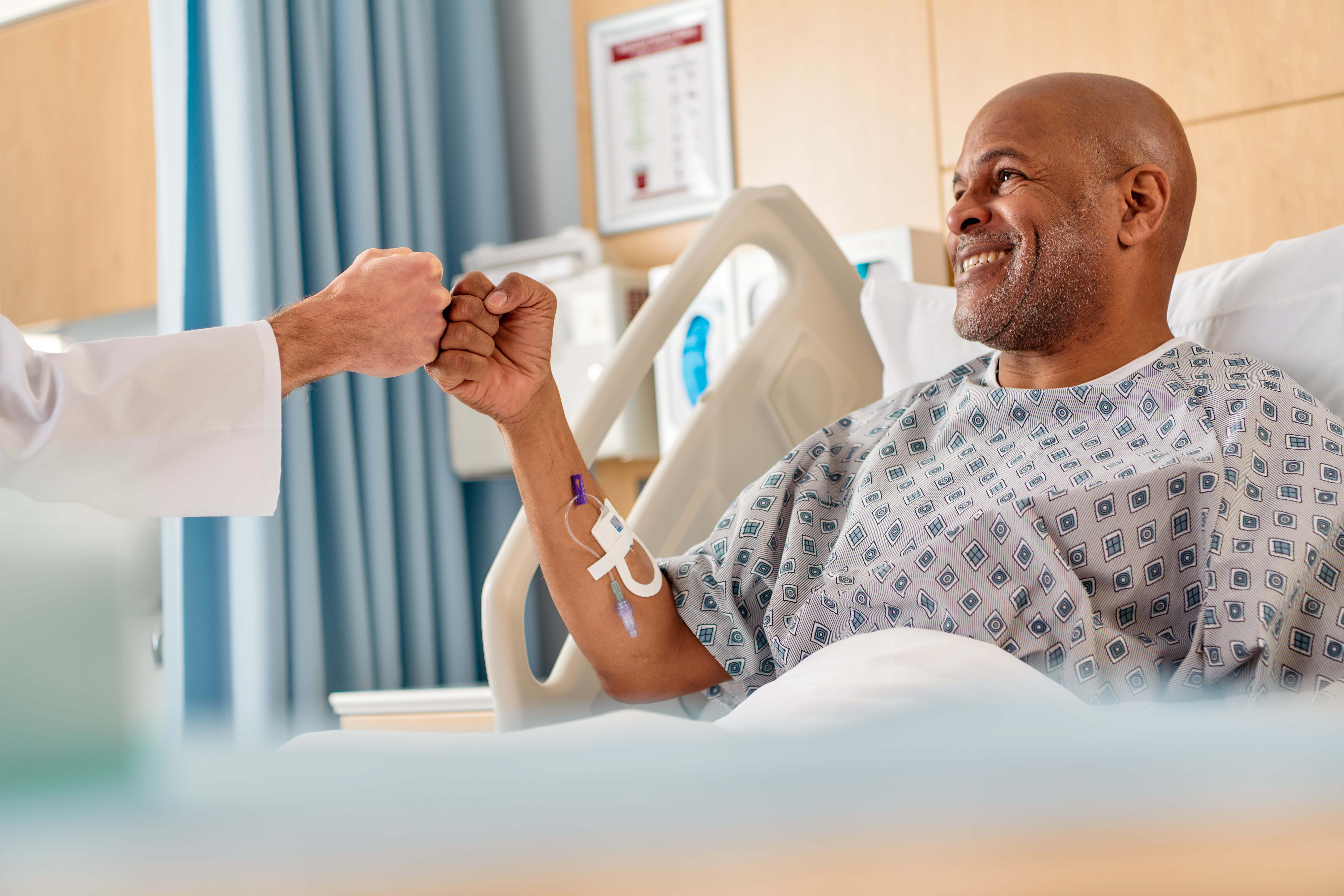Patient in a hospital bed has the Clik-FIX Catheter Securement Device on his arm as he fistbumps a healthcare provider.
